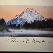 Snow-covered mountain with orange sky at sunrise, mist over a river with a solitary elk, surrounded by forest